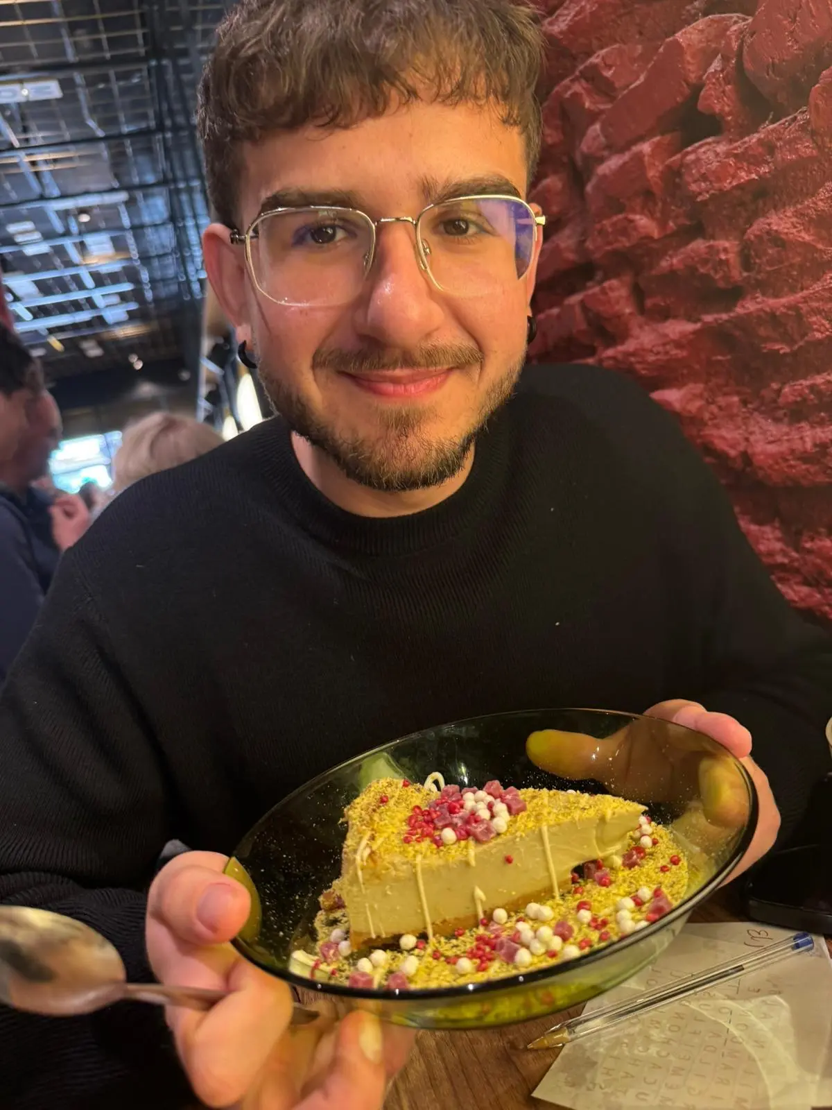 Matías smiling as he holds up a plate with a slice of cheesecake topped with pistachio