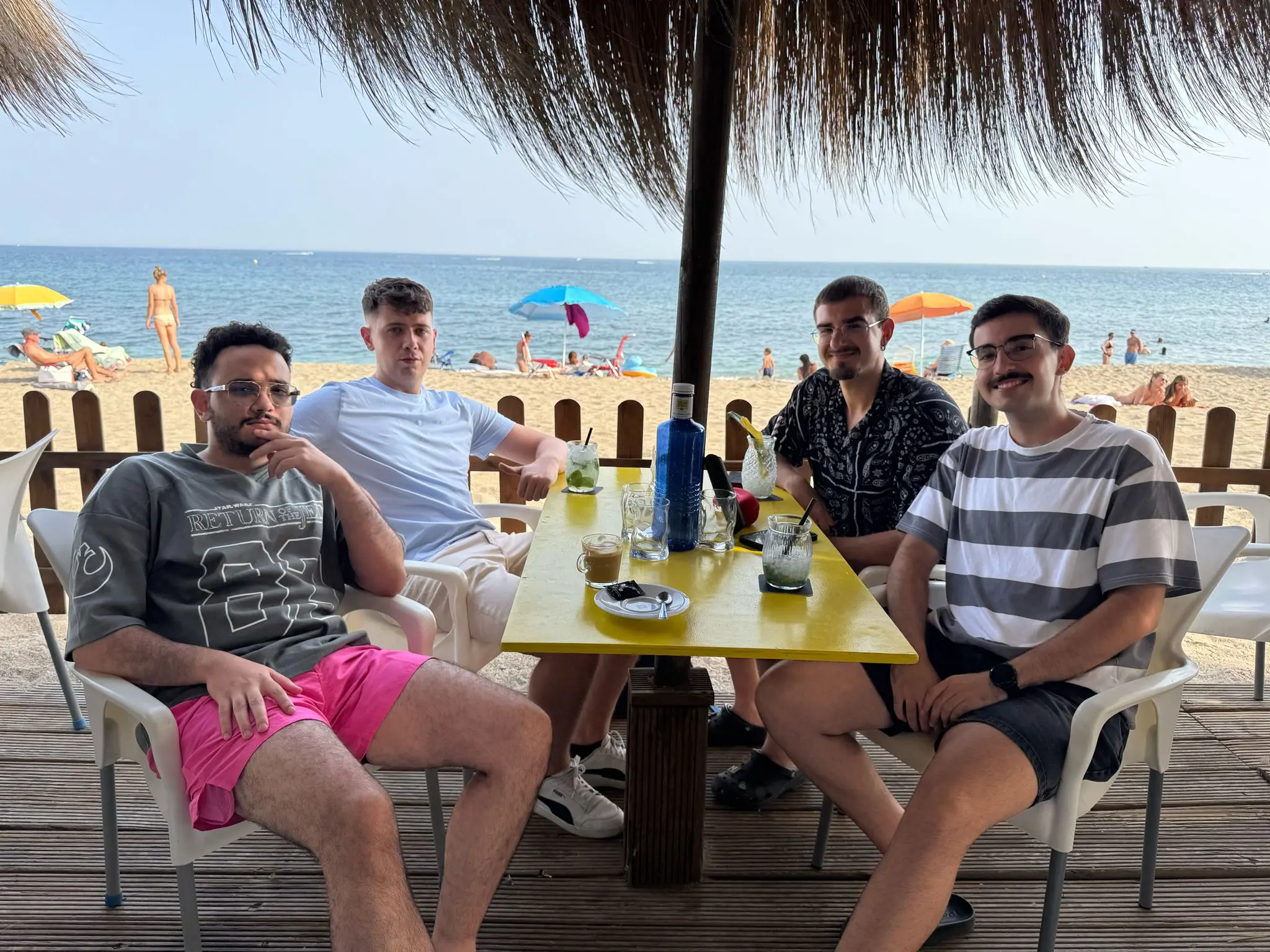 Four friends around a table at a beach bar, under a straw umbrella by the sea